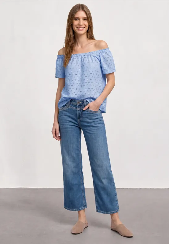 Vrouw in lichtblauwe off-shoulder blouse, culotte-jeans en beige ballerina's voor neutrale achtergrond.
