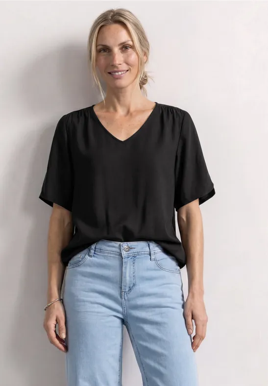 Frau in schwarzem V-Ausschnitt-Shirt und hellblauen Jeans vor weißem Hintergrund.