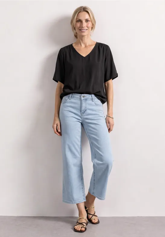 Frau in schwarzem V-Ausschnitt-T-Shirt, hellblauer Jeanshose und schwarzen Sandalen, vor neutraler Wand.