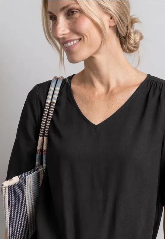 Eine lächelnde Frau in schwarzem V-Ausschnitt-Shirt trägt eine geflochtene Umhängetasche.