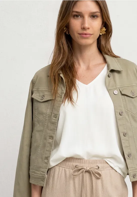 Frau in weißer V-Neck-Bluse mit olivgrüner, kurzer Jeansjacke und beige Bundfaltenhose mit Bindegürtel.