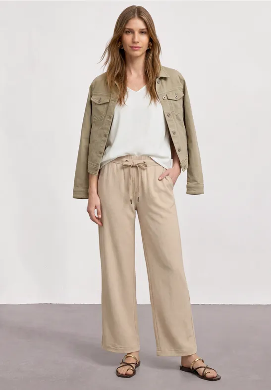 Frau in beigen, weiten Hosen, weißem T-Shirt, grünem Overshirt und flachen Sandalen.
