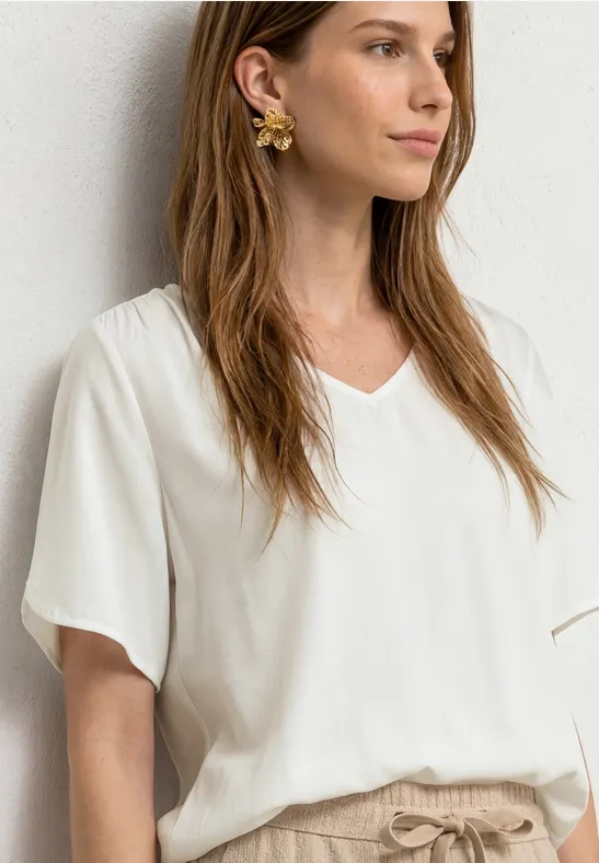 Frau in weißer V-Ausschnitt-Bluse mit kurzen Ärmeln und beiger Hose, goldene Ohrringe.