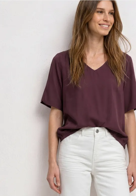 Een persoon draagt een bordeauxrood t-shirt en witte jeans voor een lichte achtergrond.