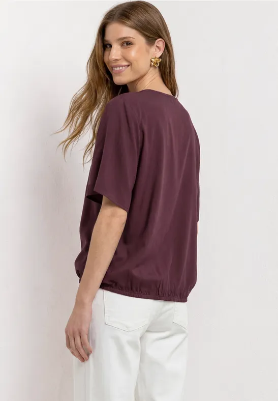 Vrouw gezien van achteren in donkerviolet T-shirt en witte broek, met los haar.