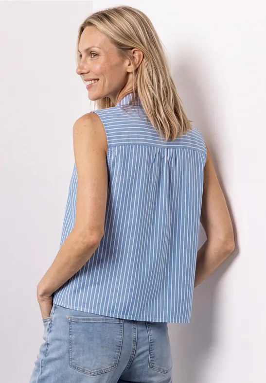 Een glimlachende vrouw van opzij en achteren gefotografeerd, in een mouwloze, lichtblauw-wit gestreepte top en jeans.