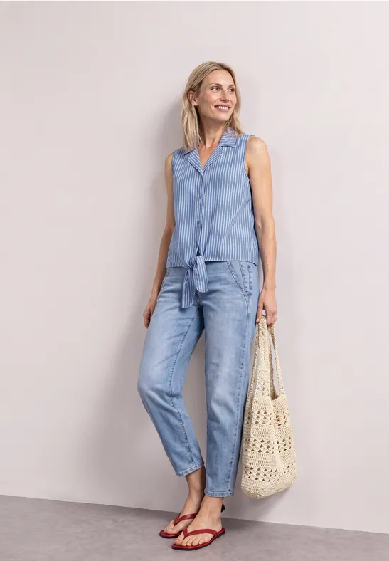 Vrouw draagt mouwloze jeanstop met knoop en spijkerbroek, rode sandalen en macramé tas voor lichte achtergrond.