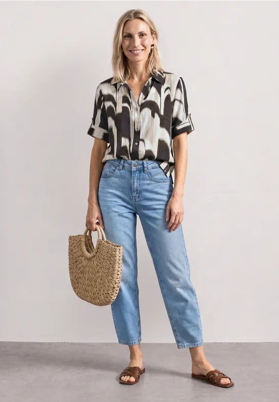 Vrouw in zwart-wit blouse, blauwe spijkerbroek en bruine sandalen, met strooien tas voor een witte achtergrond.