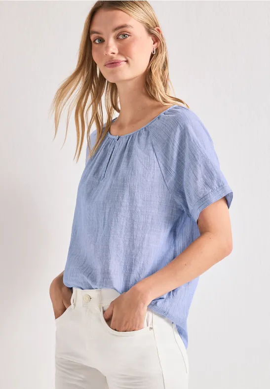 Vrouw met lichtblauw losvallend shirt en witte broek, handen in de zakken, tegen witte achtergrond.