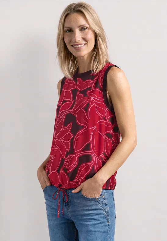 Vrouw met blond haar draagt een rood bloes zonder mouwen en blauwe jeans, handen in haar zakken.