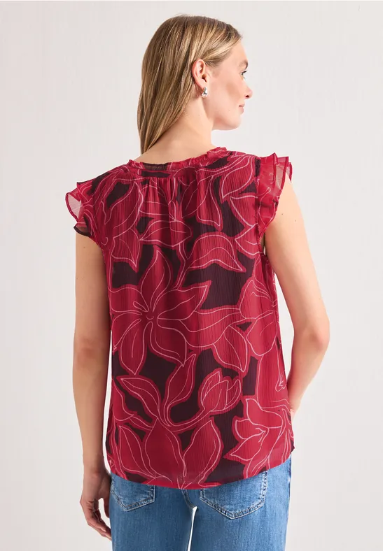 Vrouw van achteren in een mouwloze rode top met groot bladpatroon en een blauwe spijkerbroek.