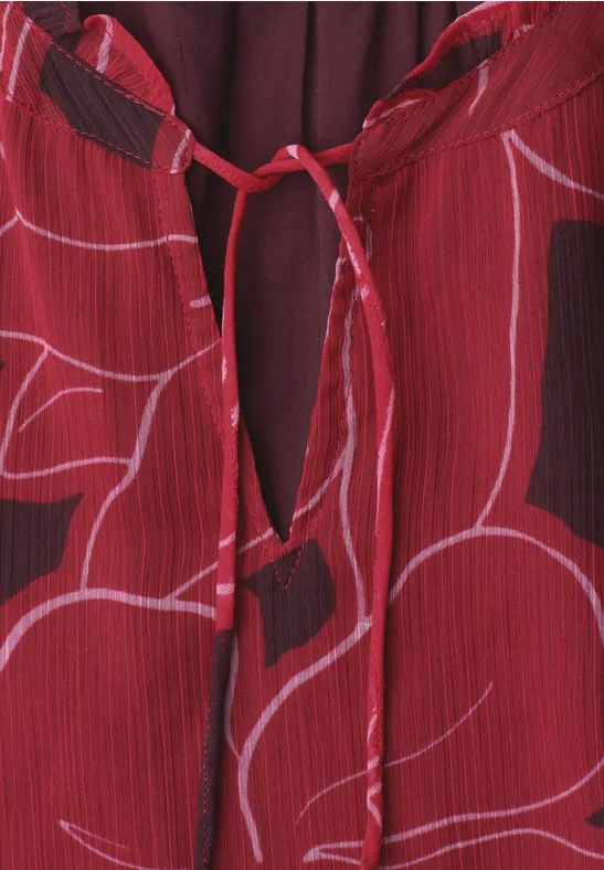Close-up van een rood kledingstuk met zachtroze bloemenprint en klein oogje aan de hals met strikje.