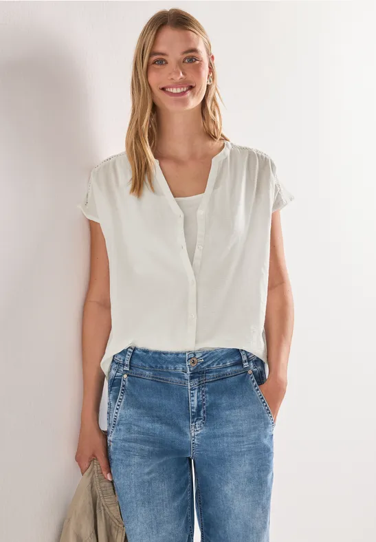 Frau mit weißer Bluse und Jeans, steht vor heller Wand mit Händen in den Hosentaschen.