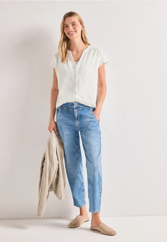 Eine Frau in weißer Bluse, blauen Jeans und Loafers hält eine helle Jacke in der Hand.