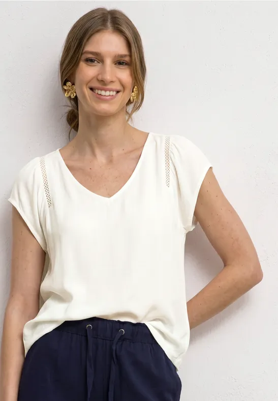 Frau in weißem V-Ausschnitt-Shirt und dunkelblauer Hose, steht lässig mit einer Hand in der Tasche.