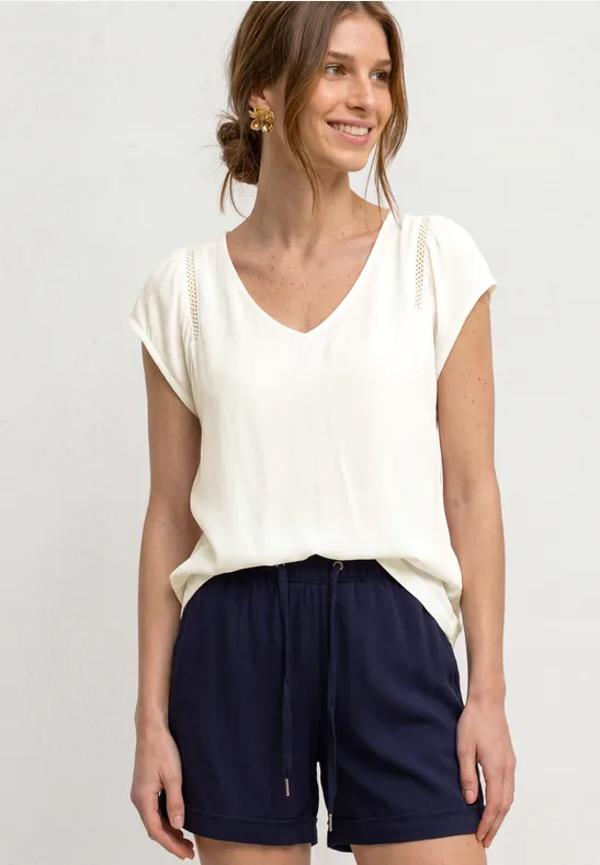 Frau in weißem V-Ausschnitt-Shirt und dunkelblauen Shorts mit Kordel, stehend vor neutralem Hintergrund.