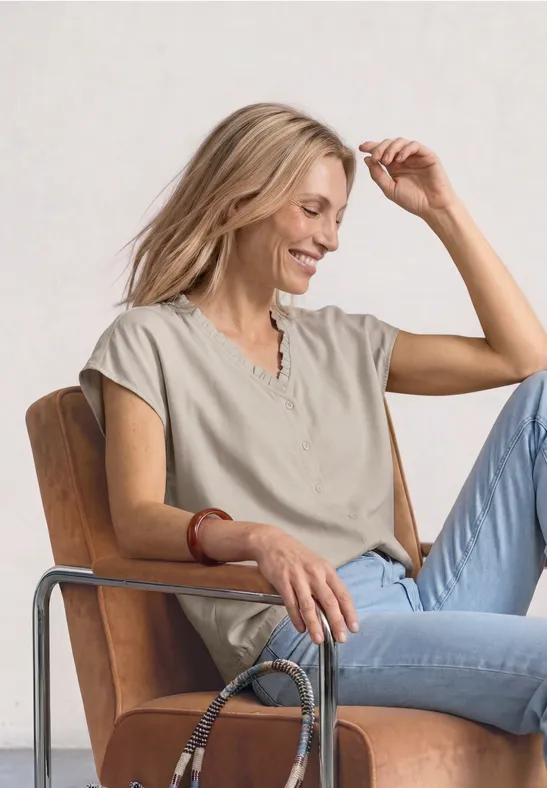 Vrouw in beige t-shirt en spijkerbroek zit ontspannen op een stoel, tas naast haar arm.