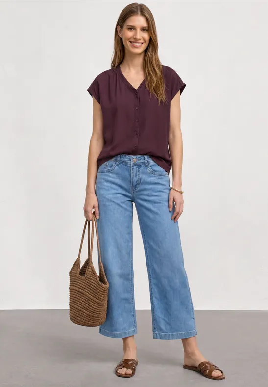 Vrouw in bordeauxrood T-shirt, wijde blauwe jeans, gevlochten tas en platte sandalen voor witte achtergrond.