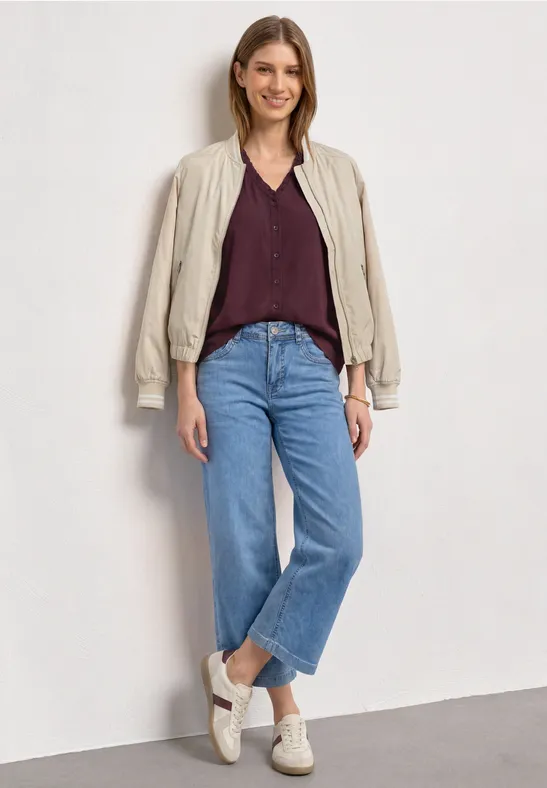 Een glimlachende vrouw in een blauwe jeans, bordeauxblouse, licht jasje en witte sneakers voor een neutrale muur.