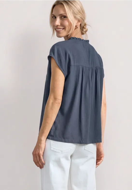 Vrouw van achteren, donkerblauw hemd met korte mouwen en witte broek, lichte achtergrond.