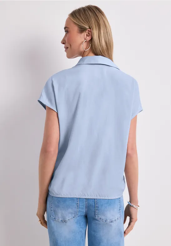 Vrouw van achteren in lichtblauwe blouse met korte mouwen en spijkerbroek voor neutrale achtergrond.