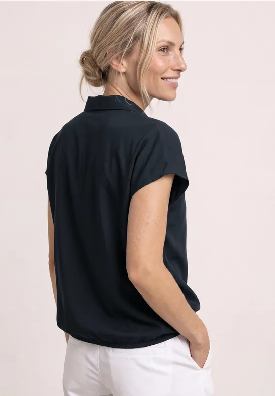 Achteraanzicht van een vrouw in een zwarte korte mouw blouse met kraag en lichte broek, handen in de zakken.