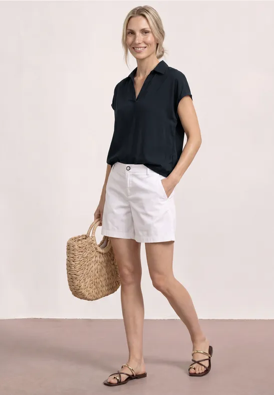 Vrouw in zwarte blouse, witte shorts, rieten tas en zwarte sandaaltjes tegen neutrale achtergrond.
