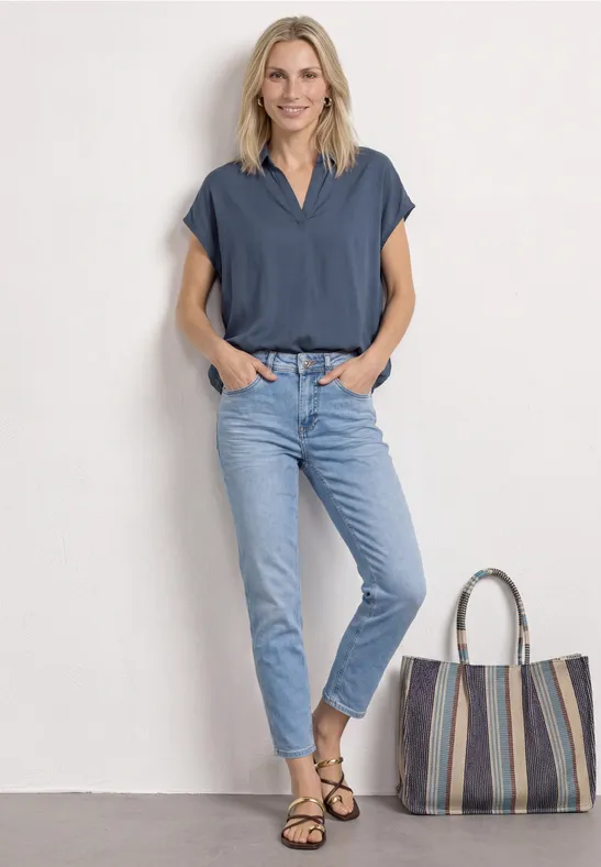 Vrouw met onscherp gezicht, draagt een blauw topje en lichtspijkerbroek, staat naast een gestreepte tas.