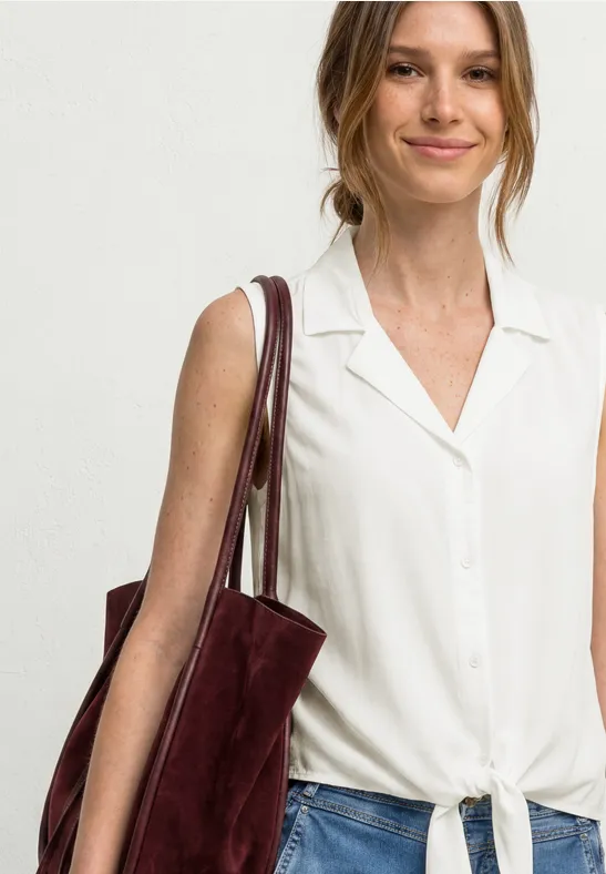 Femme en chemisier sans manches blanc noué, portant un cabas en daim bordeaux à l’épaule.