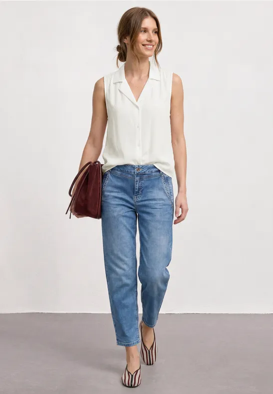Femme en chemisier blanc sans manches, jean bleu, sandales et sac rouge.