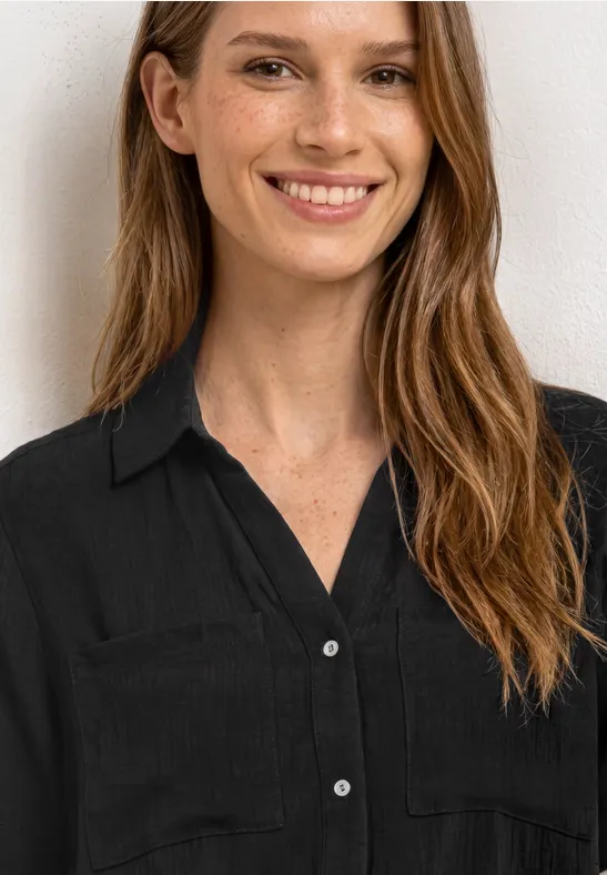 Eine Person mit langen Haaren trägt eine schwarze Bluse vor hellem Hintergrund