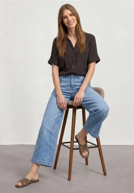 Vrouw op hoge kruk, bruine blouse, wijd jeans en sandalen voor een witte achtergrond.