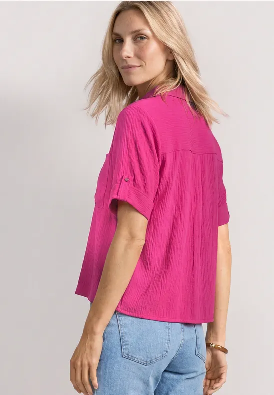 Blonde vrouw met felroze kort mouw shirt en spijkerbroek, half naar de camera gedraaid.