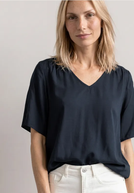 Vrouw met donkerblauw V-hals T-shirt met korte mouwen en lichte broek voor een neutrale achtergrond.