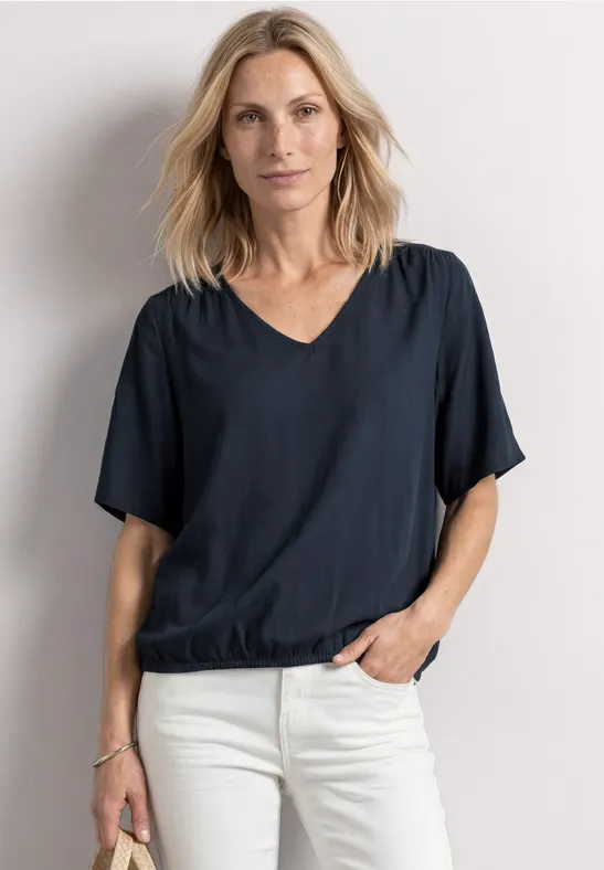 Blonde vrouw in donkerblauw V-shirt en witte broek, staand voor een lichte muur met één hand in haar zak.