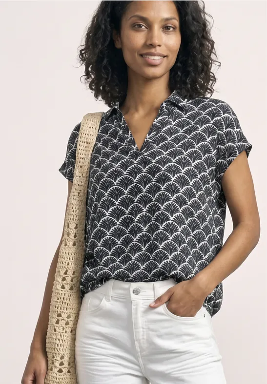 Een persoon draagt een zwarte blouse met korte mouwen en schelpmotief, witte jeans en een gehaakte strozak.