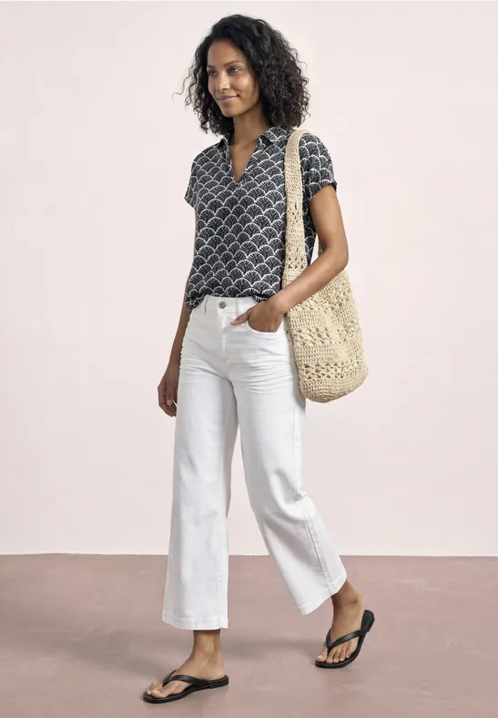 Vrouw in kort motief topje, witte cropped jeans, zwarte slippers en een rieten schoudertas.