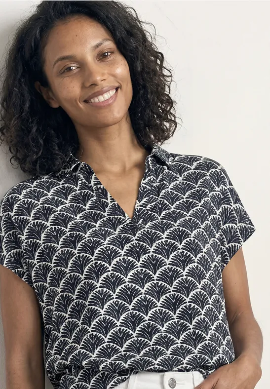 Vrouw met krullend haar draagt een zwart shirt met korte mouwen en een wit waaierpatroon.