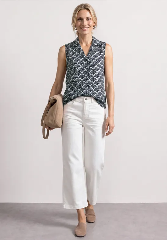 Vrouw met mouwloze printblouse, witte wijde cropped jeans, beige pumps en handtas.