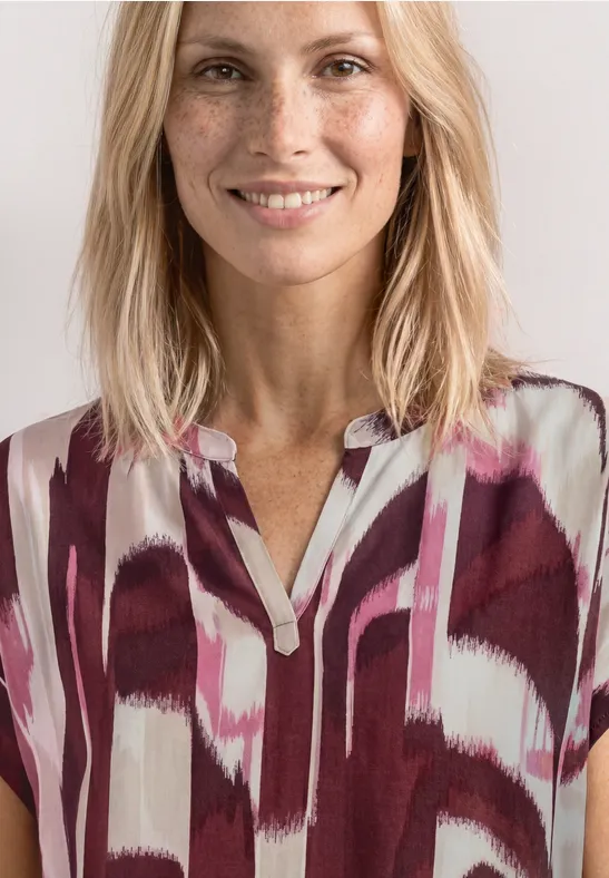 Een vrouw draagt een bordeaux-wit-roze blouse met V-hals en abstract patroon.
