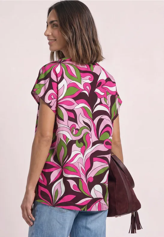 Femme porte un haut à manches courtes floral rose et vert, tenant un sac à main bordeaux.