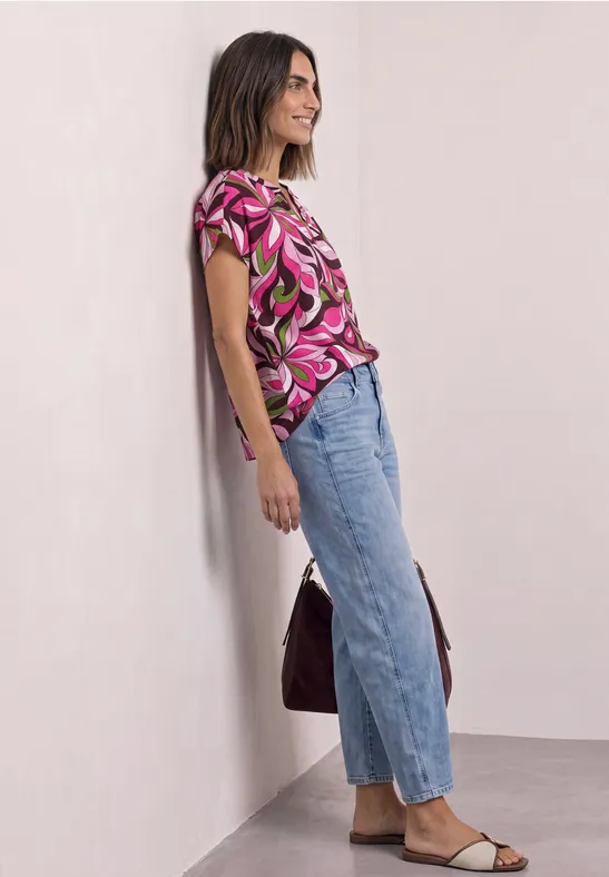 Femme appuyée contre un mur, t-shirt rose à motifs, jean bleu, sandales et sac.