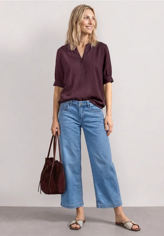 Vrouw draagt wijde lichtblauwe jeans, donkerpaarse blouse, zwarte sandalen en een bordeauxrode tas.