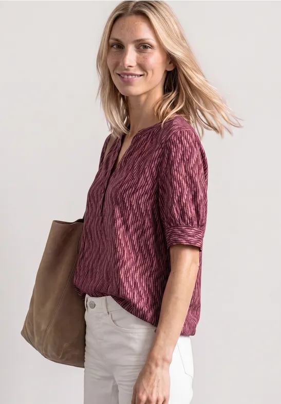 Frau in violettem Oberteil und weißen Hosen mit großer Tasche vor hellem Hintergrund.