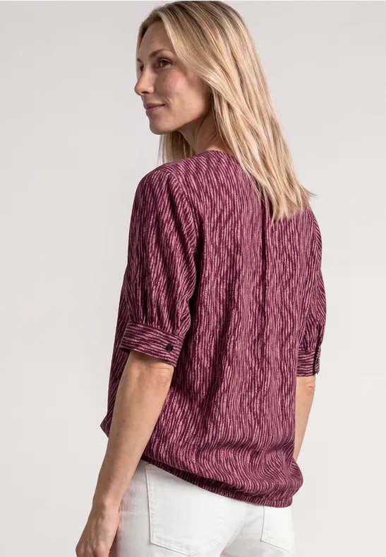 Frau mit blonden Haaren, lila Zopfmuster-Bluse und weißen Hosen, von hinten vor hellem Hintergrund.