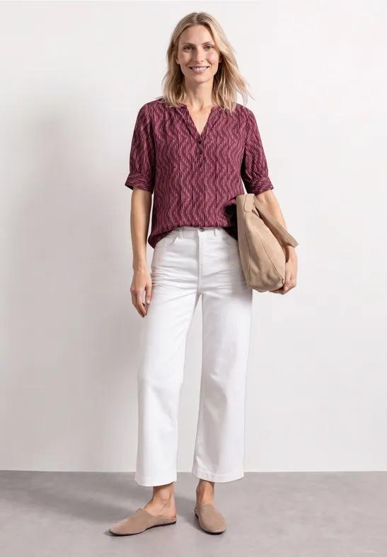 Frau in bordeauxgemustertem Oberteil, weißen Culottes, beige Slippern und mit einer beigen Tasche.