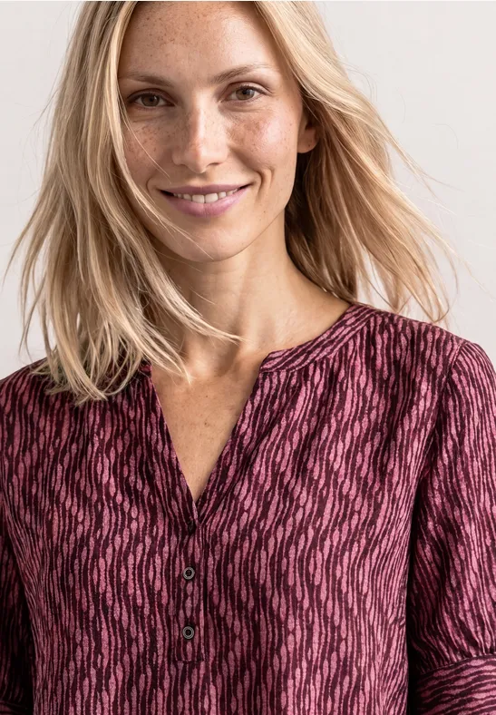 Eine Person mit langen blonden Haaren trägt eine rosé-gemusterte Bluse mit Knopfleiste.