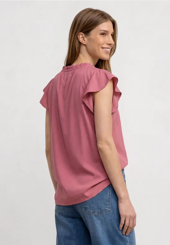 Femme portant un haut rose à manches volantées et un jean bleu, vue de côté, souriante.