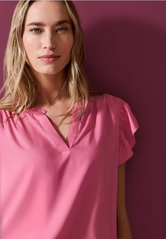 Une femme blonde en chemisier rose devant un mur rose foncé.