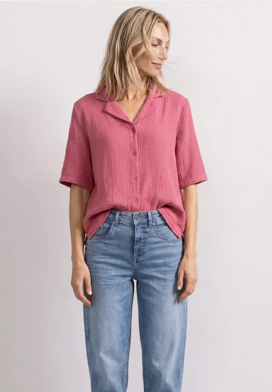 Une femme porte une chemise rose et un jean bleu devant un mur blanc.